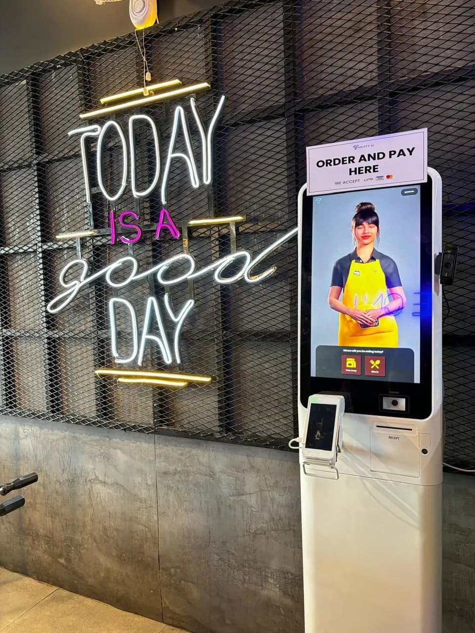 Order-and-pay kiosk with a Thrifty AI host on screen beside a neon sign in a café